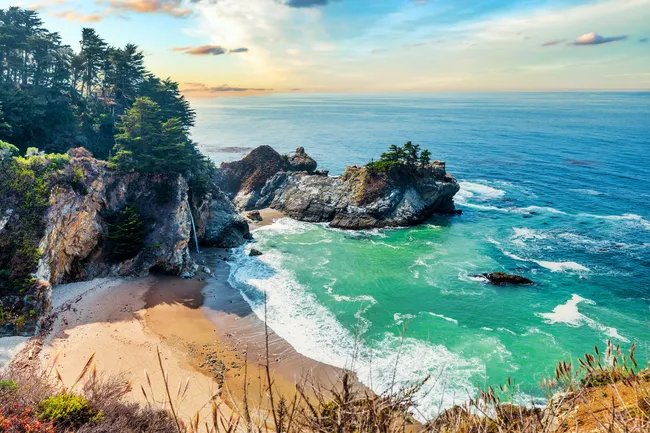 McWay Falls cascades from a rocky cliff onto a secluded beach along the Big Sur coastline, where turquoise waves wash against sea stacks and forested bluffs under a calm evening sky.