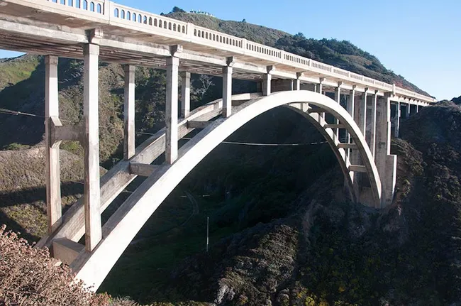 A graceful concrete arch bridge spans a steep coastal canyon, carrying a roadway high above the rugged landscape below. The bridge’s sweeping curve and tall support columns stand out against the rocky cliffs and green hillsides of the surrounding coastline.