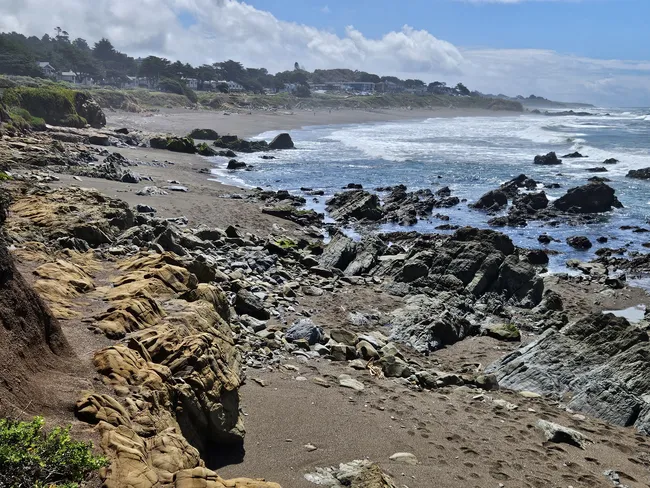 Rocky shoreline at Moonstone Beach with layered sandstone formations, scattered tide-worn boulders, gentle waves along the coast, and a distant bluff lined with trees and seaside homes under a partly cloudy sky.