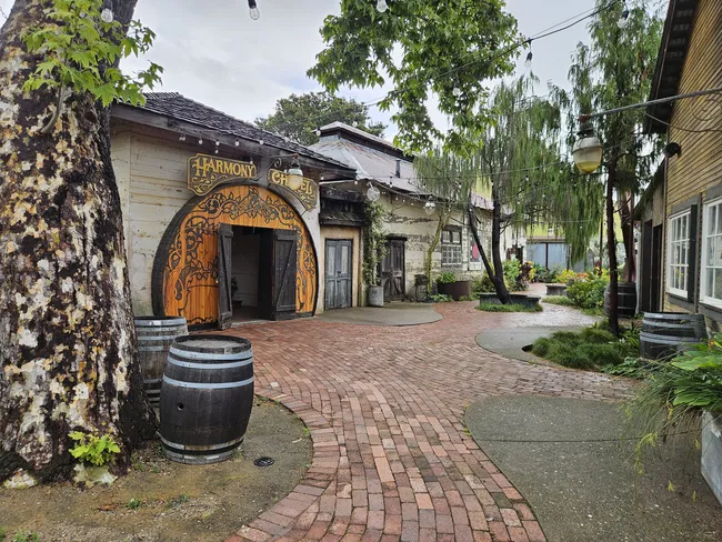 Historic buildings line a brick-paved courtyard in the tiny town of Harmony, California, featuring the ornate entrance to the Harmony Chapel, string lights overhead, mature trees, and wine barrels along a winding walkway.