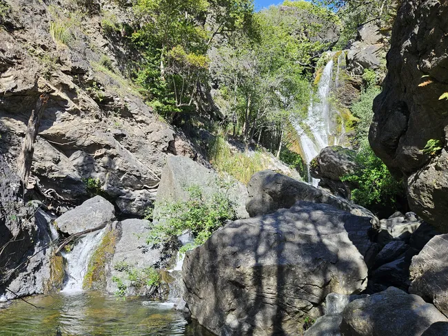 A narrow waterfall cascades down a rocky canyon wall into a clear pool, surrounded by large boulders and lush green vegetation. Sunlight filters into the sheltered gorge, highlighting the moss-covered rocks and creating a peaceful oasis within the rugged landscape.
