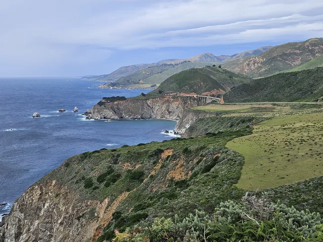 A sweeping view of the Big Sur coastline showcases rugged cliffs, rolling green hills, and deep blue ocean stretching toward the horizon. A winding coastal highway traces the shoreline, passing dramatic headlands and coves beneath a sky filled with layered clouds.