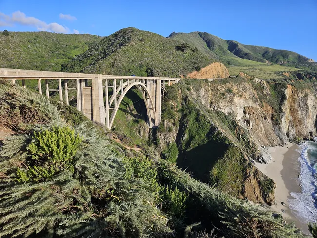 A dramatic concrete arch bridge spans a deep coastal canyon, connecting steep green hillsides high above the Pacific shoreline. Rugged cliffs descend to a narrow sandy beach below, while the bridge’s elegant design stands out against the sweeping Big Sur landscape and clear blue sky.