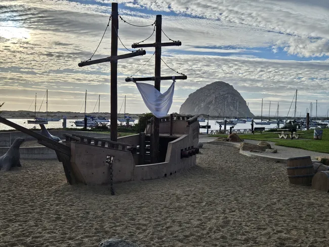 Alt text: A wooden pirate ship playground sits in a sandy play area at Tidelands Park in Morro Bay, with sailboats moored in the harbor and Morro Rock rising in the background beneath a dramatic cloud-filled sky.