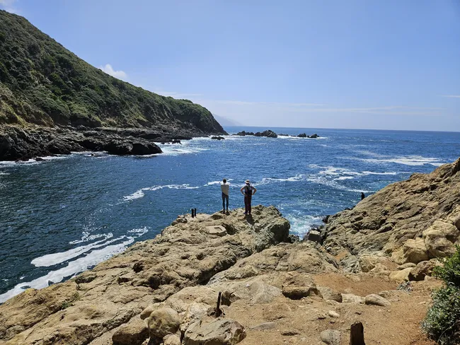 A rocky coastal cove opens to the Pacific Ocean, where two visitors stand on a large outcrop taking in the expansive seaside view. Steep green hills and rugged cliffs frame the sheltered inlet, while gentle waves swirl across the deep blue water below.