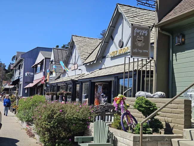 Storefronts and boutique shops line a sunny sidewalk in downtown Cambria, featuring charming coastal architecture, colorful landscaping, outdoor seating, and pedestrians strolling beneath a clear blue sky.