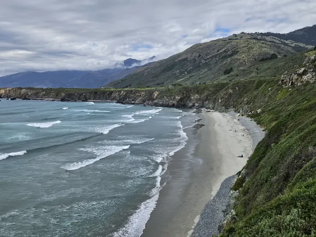 A long crescent-shaped beach stretches beneath steep coastal bluffs, with gentle waves rolling onto the shore from the Pacific Ocean. Rugged green mountains rise behind the coastline, while low clouds drift across the sky above the expansive Big Sur landscape.