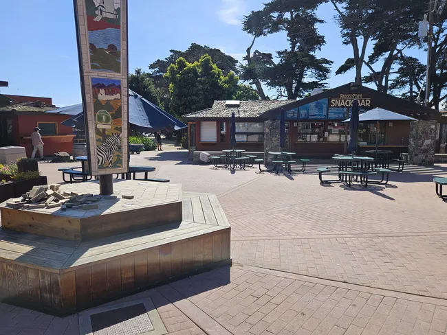 Open-air plaza at Ragged Point on California’s Highway 1, featuring picnic tables, a snack bar, a decorative pillar with coastal artwork and a Highway 1 emblem, and tall cypress trees under a clear blue sky.