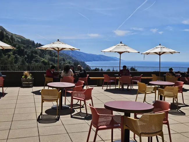 An outdoor terrace overlooks the dramatic Big Sur coastline, with colorful tables and chairs arranged beneath large white umbrellas. Visitors relax and dine while enjoying sweeping views of forested mountains, rugged cliffs, and the Pacific Ocean under a clear blue sky.