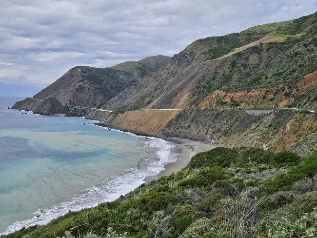 A dramatic stretch of coastline features steep mountainsides descending to a secluded beach and pale turquoise waters below. A winding highway and an arched coastal bridge trace the rugged shoreline, while clouds hang over the expansive Big Sur landscape.