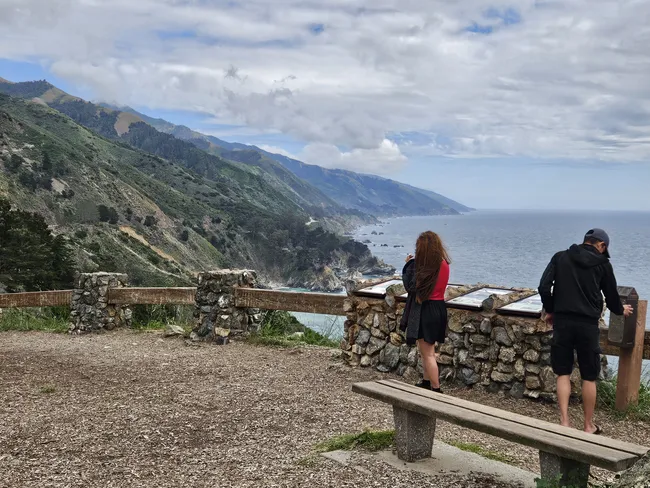 A scenic overlook features visitors standing beside a stone viewing wall, taking in panoramic views of the rugged Big Sur coastline. Steep mountains and rocky cliffs descend to the Pacific Ocean below, while low clouds drift across the sky above the dramatic landscape.