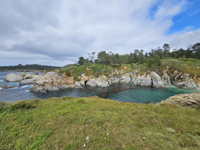 A small turquoise cove is surrounded by rugged white rock formations and coastal vegetation, creating a sheltered inlet along the shoreline. Pine-covered bluffs rise above the water, while dramatic clouds drift across the sky over the scenic coastal landscape.