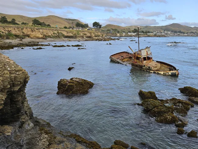 A rusted shipwreck rests in shallow coastal waters off Estero Bluffs, surrounded by rocky shoreline and calm ocean waves, with rolling hills and a small seaside town in the background beneath a cloudy sky.
