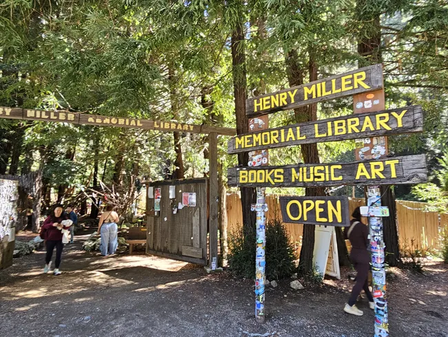 A rustic wooden sign marks the entrance to the Henry Miller Memorial Library, nestled among towering redwood trees in a forested setting. Visitors walk through the shaded grounds beneath signs advertising books, music, and art, creating a welcoming atmosphere for exploration and creativity.