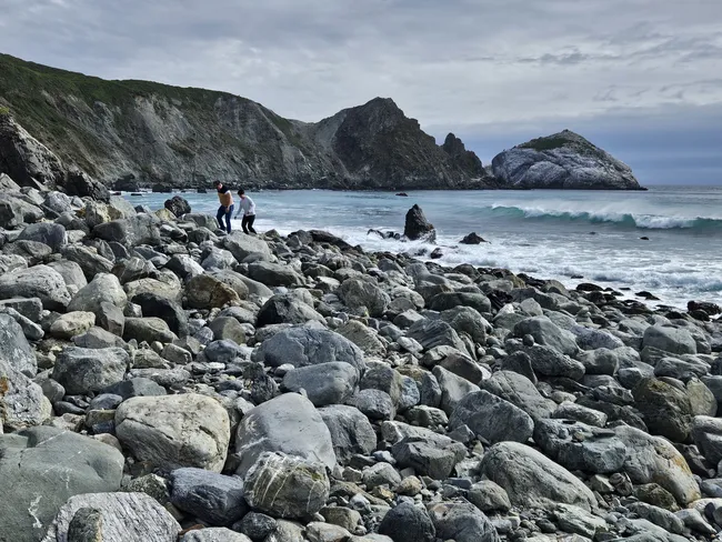 A rocky shoreline is covered with smooth gray boulders, where visitors carefully explore the coast beside crashing waves. Steep cliffs and rugged offshore rock formations frame the cove, while the ocean stretches beneath a moody, cloud-filled sky.