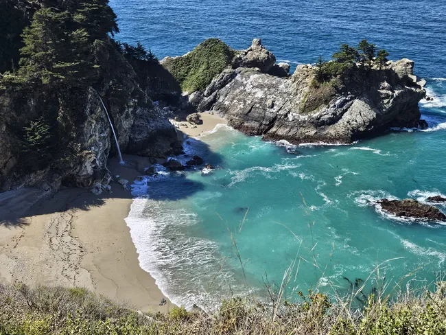 A slender waterfall cascades directly from a coastal cliff onto a secluded sandy beach, creating one of the most iconic views along the Big Sur coast. Turquoise waters surround rocky offshore islets and rugged cliffs, framing the picturesque cove beneath a clear sky.