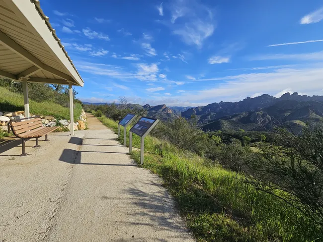 Scenic overlook trail with a bench, interpretive signs, and a covered shelter, offering panoramic views of rugged mountain peaks and green valleys beneath a bright blue sky.