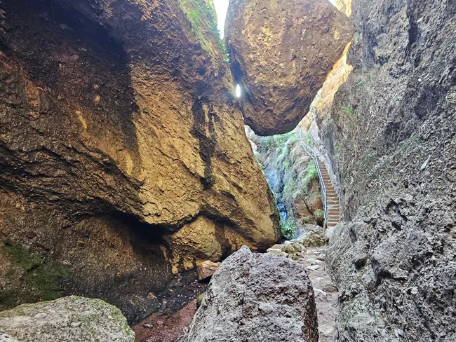 Narrow rock passage in Bear Gulch Cave with towering canyon walls, a large boulder wedged overhead, and a steep staircase with a handrail leading upward through the rocky corridor.