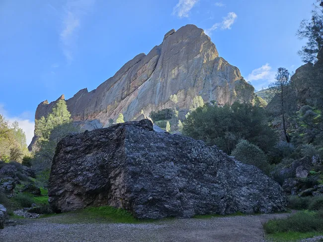 A massive rock formation rises above a large boulder in a rugged canyon landscape, with scattered trees and shrubs beneath a clear blue sky.