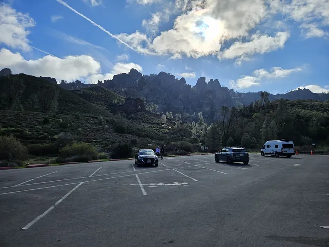 Parking area at a trailhead with a few vehicles parked in front of rugged rock formations and rolling hills, under a bright sky with scattered clouds.