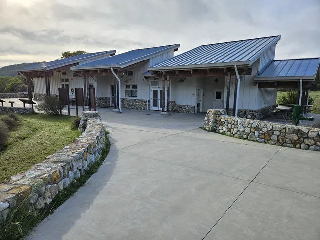 Modern visitor center building with metal roofs, stone accents, and covered entrances, connected by a wide concrete walkway and low stone walls in a natural park setting.