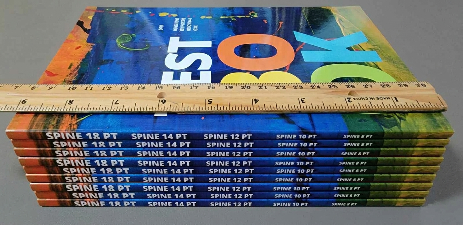 Ein Stapel identischer Bücher mit dem Titel „TEST BOOK“, auf deren oberem Einband flach ein Holzlineal liegt, um das Endformat zu messen.