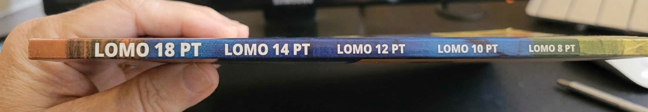 Mano sosteniendo el lomo de un libro con muestras tipográficas "LOMO" en tamaños de 18, 14, 12, 10 y 8 puntos.