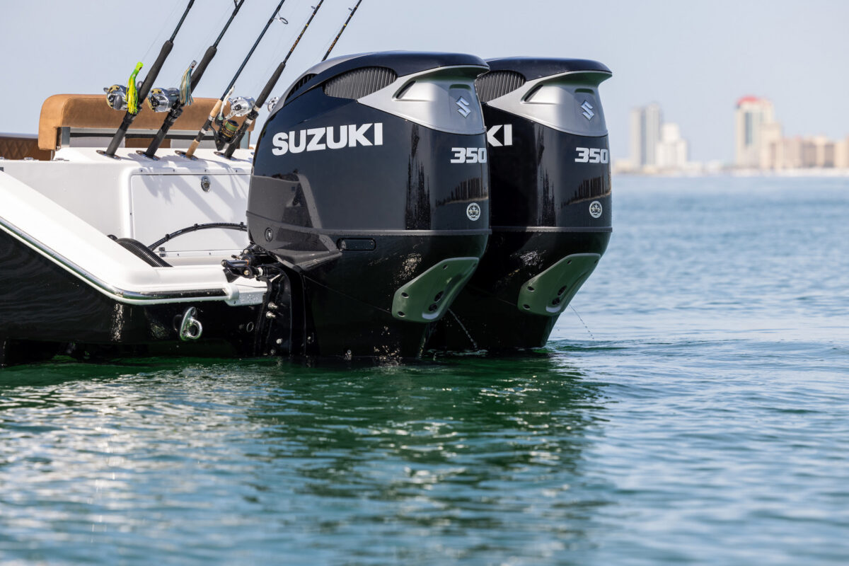 Twin Suzuki 350 outboard engines partially submerged at the stern of a fishing boat, with multiple fishing rods mounted nearby and a distant city skyline visible across the calm blue water.