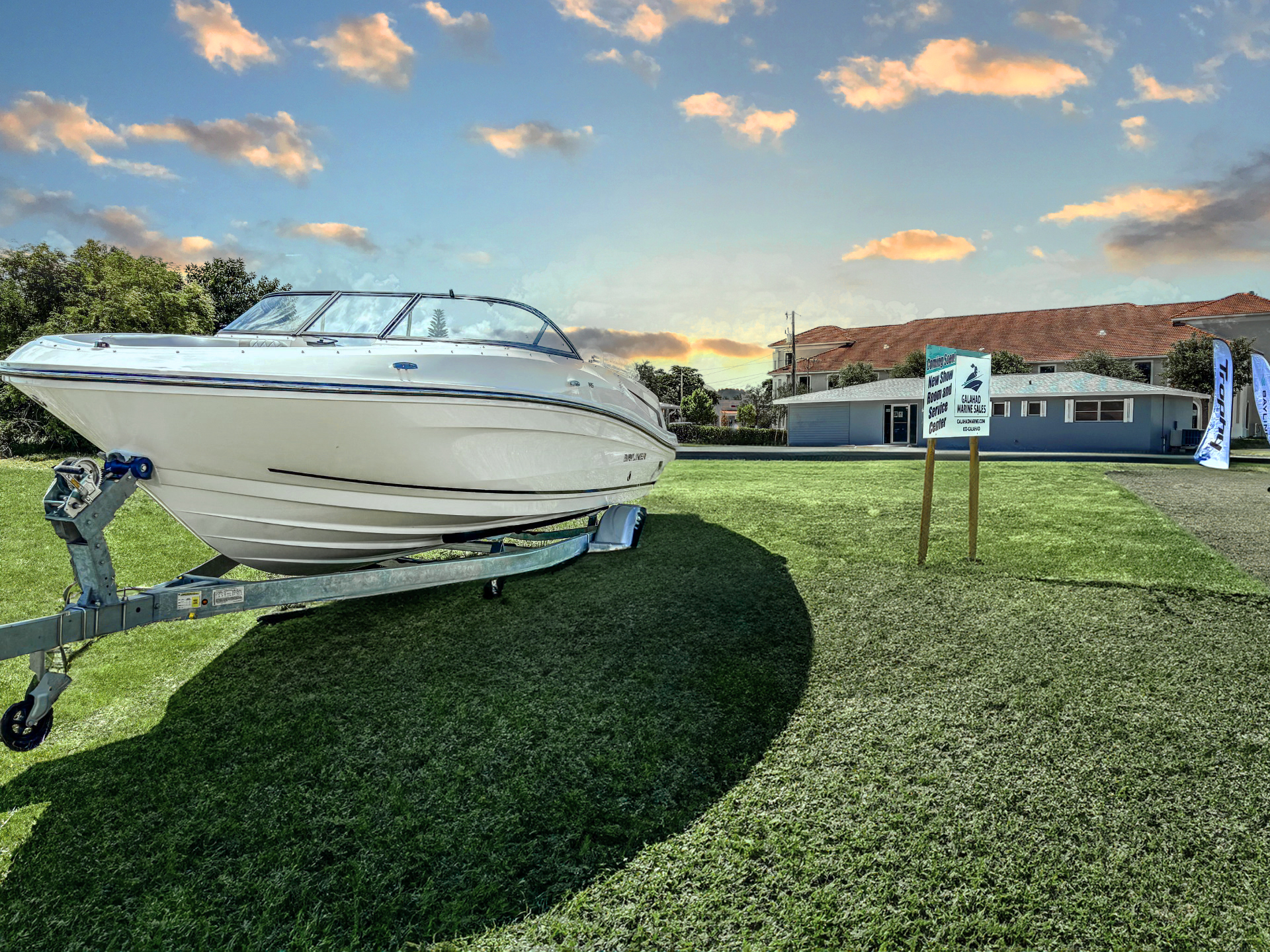White bowrider boat on a trailer displayed on a grassy lawn at sunset, with a Galahad Marine Sales sign and building in the background under a colorful sky.