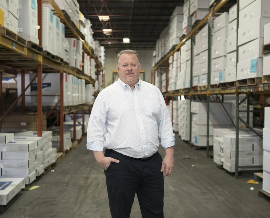 A man in a white shirt and dark pants stands in a warehouse aisle lined with stacked boxes on shelves.