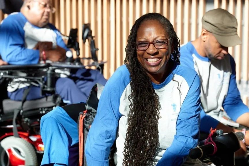 A woman with long hair and glasses smiles while sitting. Two men in similar shirts are in the background; one in a wheelchair and one looking down.