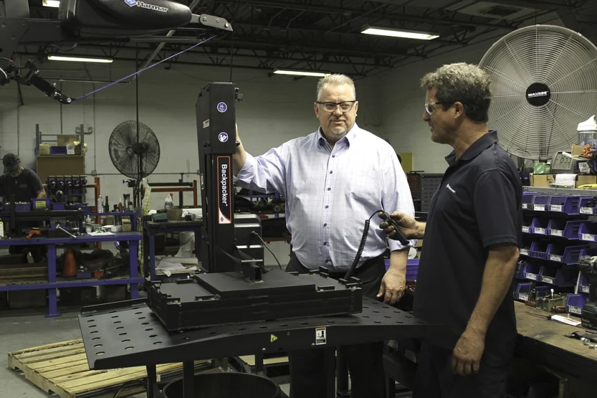 Two men in a workshop stand near industrial machinery. One man wears a light shirt and glasses, the other a dark polo shirt, holding a tool. Equipment and shelves are visible in the background.