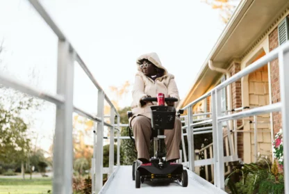 Person in a hooded jacket uses a mobility scooter to descend a ramp in front of a house.