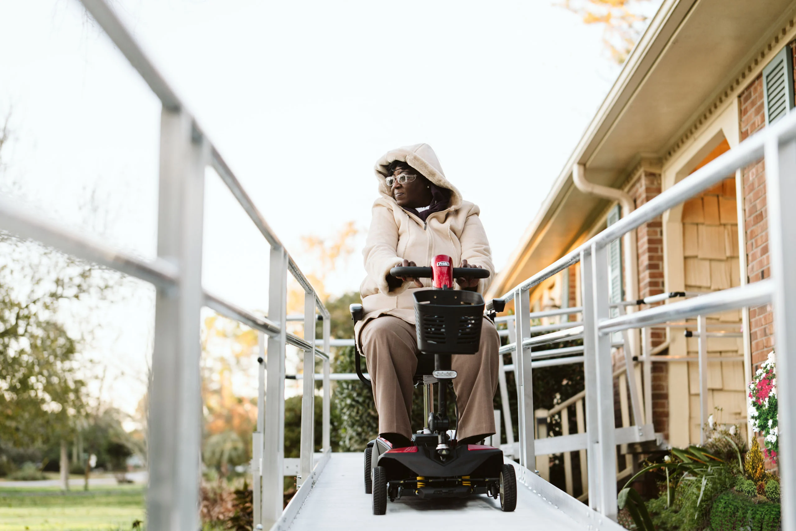 Person in a hooded jacket uses a mobility scooter to descend a ramp in front of a house.
