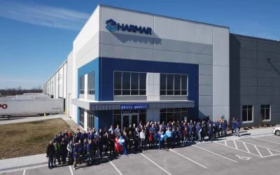 Group of people standing outside a large industrial building labeled "HARMAR" on a clear day.