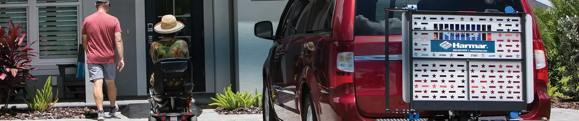 A person in a hat uses a mobility scooter to approach the entrance of a house, following another person. A red vehicle with a mobility lift is parked nearby.