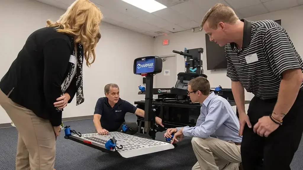 Four people examining a mechanical device with blue accents indoors, involving a standing woman and three kneeling men.
