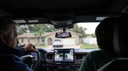 View from inside a car showing two people in front seats, dashboard display with a rear-view camera image, and a house with a parked vehicle in the driveway.