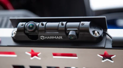 Close-up of a Harmar branded device mounted on a metal surface with red star-shaped cutouts. It features two lenses and is connected by wires.