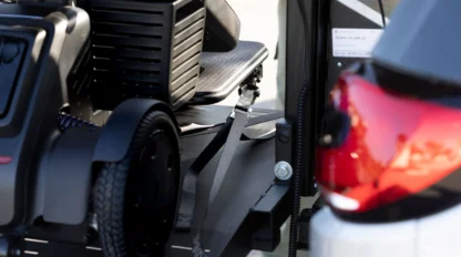 Electric scooter secured on a car hitch-mounted carrier with a strap, next to the rear of a white and red vehicle.