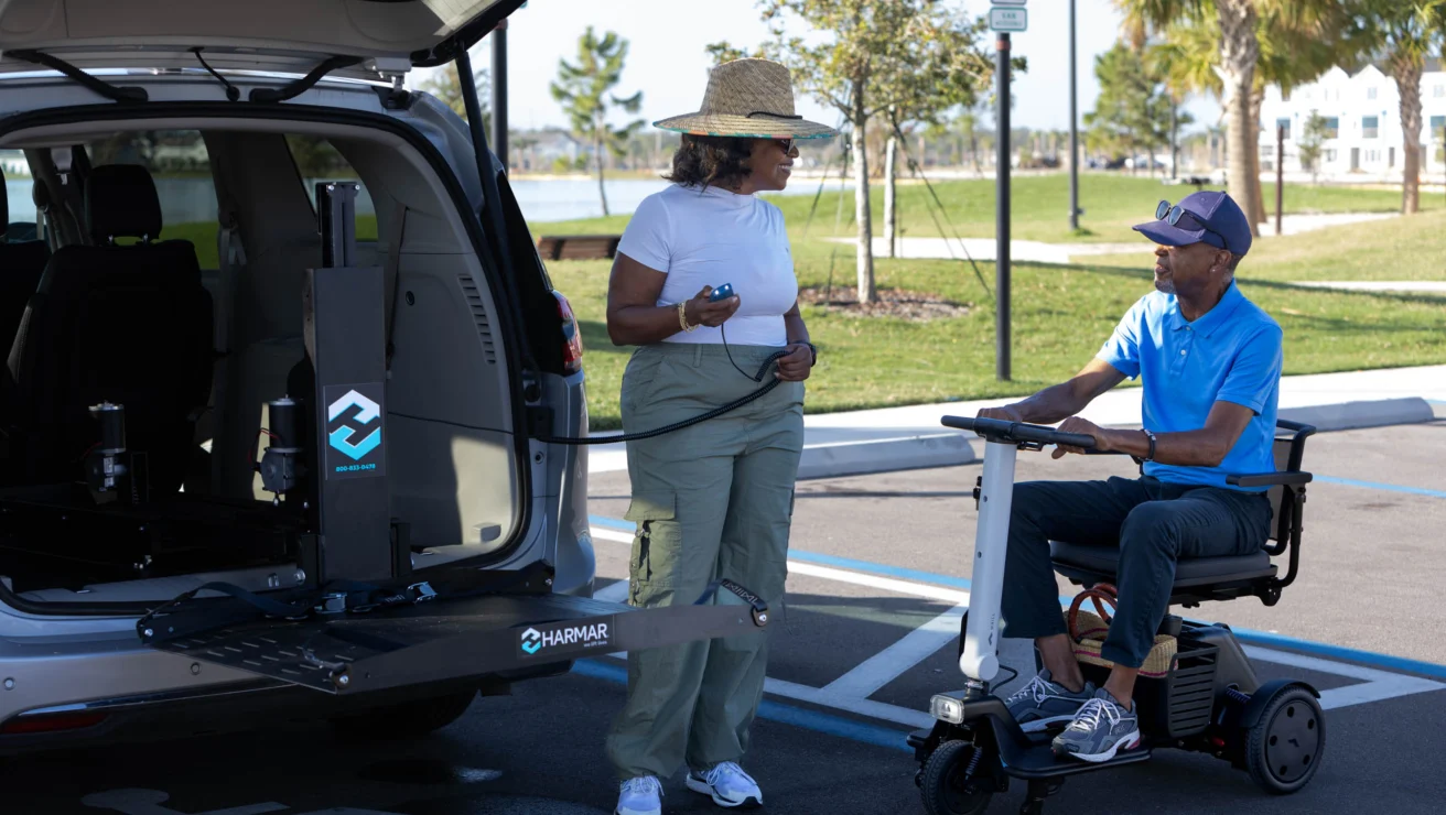 A woman helps a man in a mobility scooter near a van with an open lift. They are in a sunny parking lot with palm trees and handicap signs.