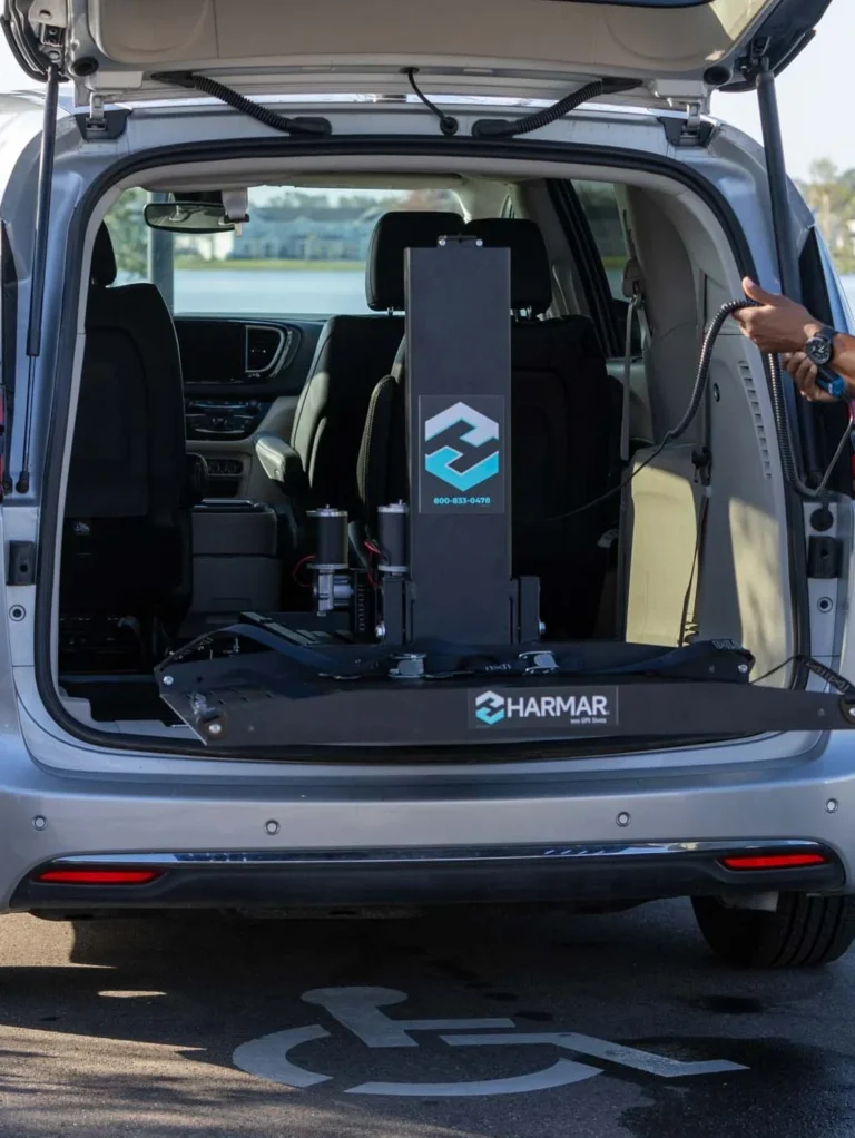 A woman operates a wheelchair lift at the back of an open minivan, while a man on a mobility scooter watches. The scene is in a parking lot by a body of water.