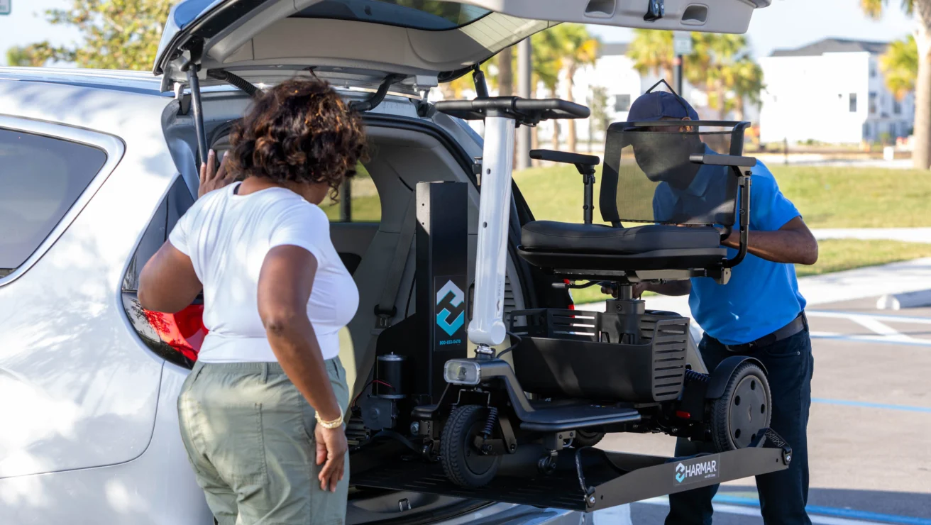 A person operates a lift to load a mobility scooter into the back of a vehicle while another person watches.