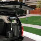 An open SUV trunk showing a Harmar lift installed inside, with a strap hanging down. A suburban house with a lawn and driveway is visible in the background.