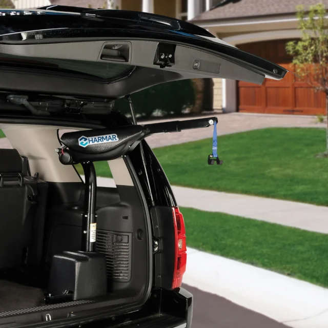 An open SUV trunk showing a Harmar lift installed inside, with a strap hanging down. A suburban house with a lawn and driveway is visible in the background.