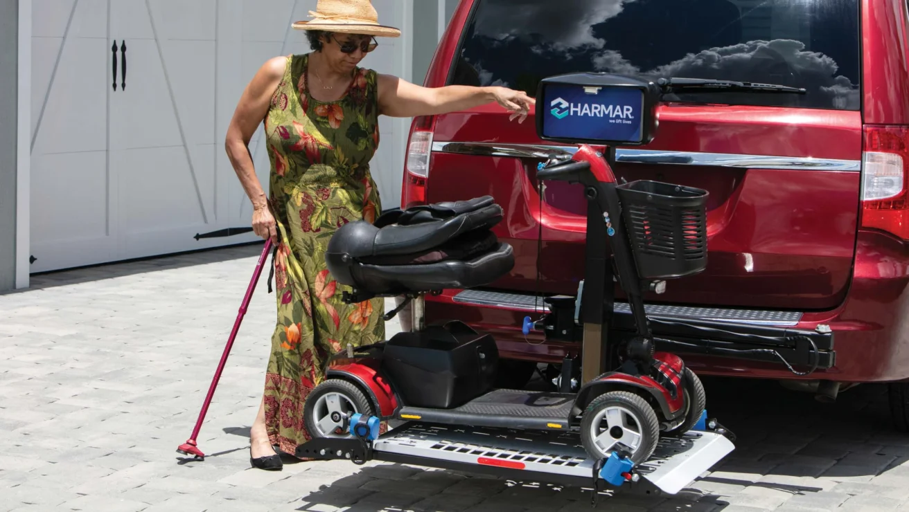 A woman in a floral dress and sun hat uses a cane and operates a mobility scooter lift attached to a red SUV in a driveway.