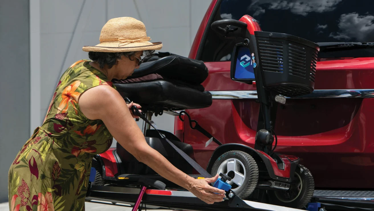 A woman in a sun hat secures a mobility scooter to the rear of a red van using a lift.