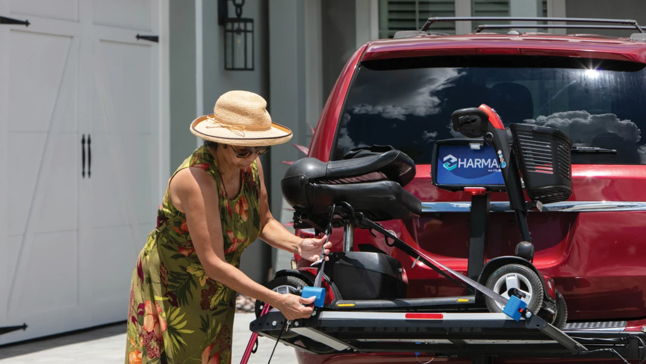 Woman in a floral dress and straw hat adjusts a mobility scooter on the back of a red SUV parked in a driveway.