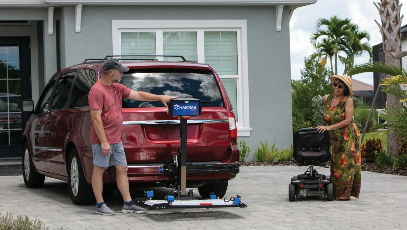 A man operates a lift on a red minivan, with a woman nearby holding a mobility scooter on a driveway outside a house.