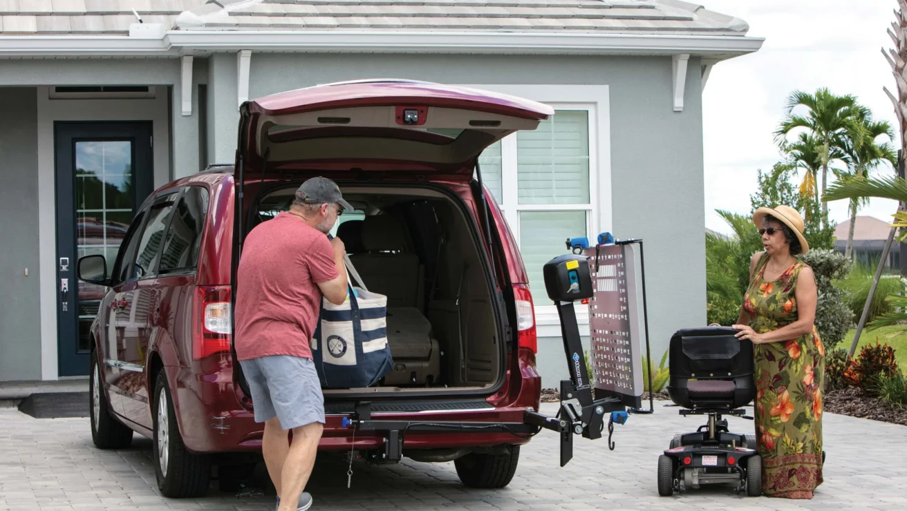 A man in a red van loads items into the back using a lift. A woman stands nearby with a mobility scooter, outside a suburban house.